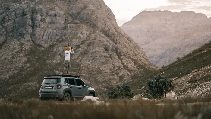 Free A man standing on an SUV roof using binoculars to view a mountain landscape. Stock Photo